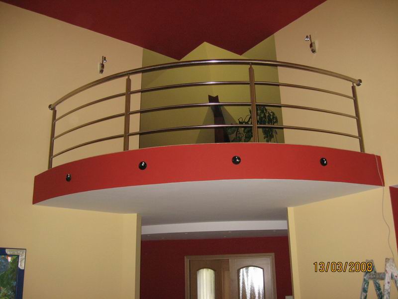 Wnętrze z antresolą zabezpieczoną metalową balustradą, pomalowane ściany w odcieniach żółci i czerwieni, widoczne oświetlenie punktowe i roślina doniczkowa na górze.